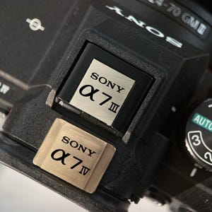 May include: Close-up of a black Sony camera body with a silver and gold finish. The camera features the "SONY α7 III" and "SONY α7 IV" logos. The lens is partially visible, along with the mode dial set to "AUTO".