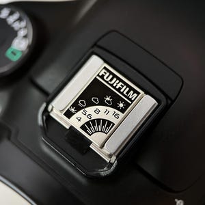 Pode incluir: Close-up de uma câmera Fujifilm preta com um medidor de luz. O medidor de luz tem uma moldura prateada e exibe o logotipo da Fujifilm e várias configurações de abertura. O mostrador da câmera é visível ao fundo.