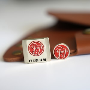 Könnte beinhalten: Zwei silberne Metallstifte mit einem roten und weißen Fuji-Logo. Der größere Stift trägt den Text "FUJIFILM" unter dem Logo.