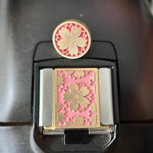 May include: A brass and pink floral design hot shoe cover and a matching round accessory. The design features a cherry blossom pattern. The accessories are set against a black background.
