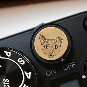 May include: A close-up of a camera with a gold-colored knob featuring a black and white line drawing of a cat's face. The camera has a black dial with the words "SCENE", "AUTO", "P", "S", and "A" printed on it. The camera also has a black switch with the words "ON" and "OFF" printed on it.