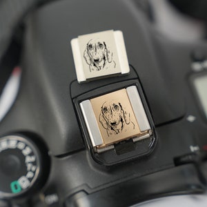 May include: A black camera with a silver flash attachment. The flash attachment has a small, engraved image of a dog on it.