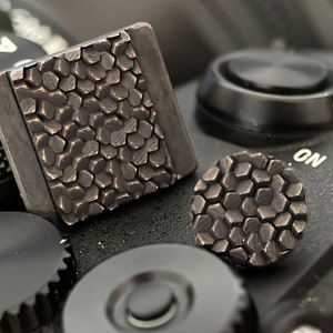 May include: Two black camera grip covers with a hexagonal pattern. The covers are made of a textured material and are designed to improve grip on a camera.