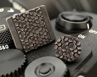 Può includere: Due copri impugnatura per fotocamera neri con un motivo esagonale. Le coperture sono realizzate in materiale testurizzato e sono progettate per migliorare la presa su una fotocamera.