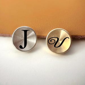 Op de afbeelding: Twee ronde metalen knopen, &eacute;&eacute;n zilverkleurig en &eacute;&eacute;n goudkleurig, elk met een zwarte letter. De zilveren knoop heeft de letter "J", en de gouden knoop heeft de letter "V". De knopen liggen op een witte ondergrond met een bruine achtergrond.