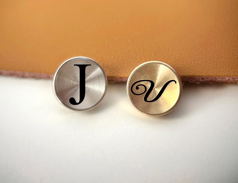 Op de afbeelding: Twee ronde metalen knopen, &eacute;&eacute;n zilverkleurig en &eacute;&eacute;n goudkleurig, elk met een zwarte letter. De zilveren knoop heeft de letter "J", en de gouden knoop heeft de letter "V". De knopen liggen op een witte ondergrond met een bruine achtergrond.
