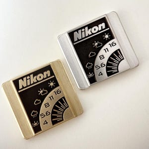 Pode incluir: Dois acessórios de fotómetro para câmaras Nikon. Um é dourado, o outro prateado. Ambos apresentam o logótipo Nikon e uma escala de tempo e luz. A escala inclui ícones de sol, nuvens e chuva, com os números 4, 5,6, 8, 11 e 16.