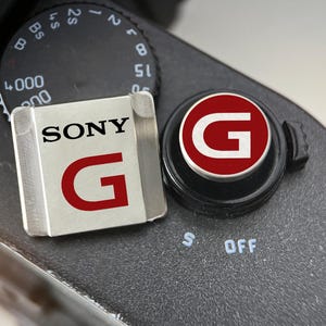 May include: Two camera accessories are shown on a black surface. One is a silver rectangle with the word "SONY" and a red "G". The other is a black cap with a red circle and a white "G".
