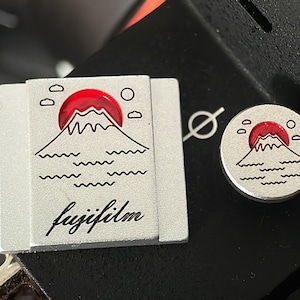 May include: A silver camera accessory with a black and white illustration of Mount Fuji, a red sun, and clouds. The text "fujifilm" is written in cursive below the illustration.