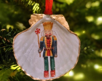 Könnte beinhalten: Ein weißer Muschelschmuck mit goldener Verzierung, der ein gemaltes Bild eines Nussknackers zeigt, der einen Zuckerstock hält. Der Nussknacker trägt eine blaue und rote Uniform mit goldenen Akzenten.