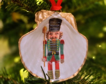 Könnte beinhalten: Ein Weihnachtsschmuck mit einem handgemalten Nussknackersoldaten in einer weißen Muschelschale. Die Schale hat einen goldfarbenen Rand und wird von einem roten Band gehalten. Der Nussknacker trägt eine grüne Jacke, eine rote Hose und einen schwarzen Hut.
