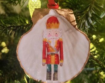 Könnte beinhalten: Ein weißer Muschelschmuck mit goldener Verzierung, der ein gemaltes Bild eines Nussknackersoldaten zeigt, der eine rot-goldene Uniform und eine Krone trägt.