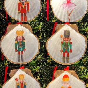 Peut inclure: Six décorations de Noël représentant des personnages de casse-noisette et de ballerine peints sur des coquillages. Les coquillages sont peints en or et ont des rubans rouges pour les suspendre. Les personnages sont habillés en costumes traditionnels de casse-noisette, avec une ballerine en robe rose.