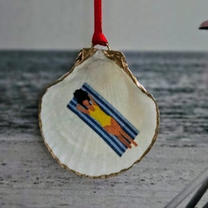 May include: A decorative oyster shell ornament with a gold-painted edge and a red ribbon for hanging. The shell features a painted scene of a person in a yellow swimsuit sunbathing on a blue and white striped towel.
