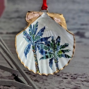 Könnte beinhalten: Ein weißer Muschelschmuck mit goldener Umrandung und einem gemalten Design aus zwei Palmen mit blauen, grünen und gelben Blättern.