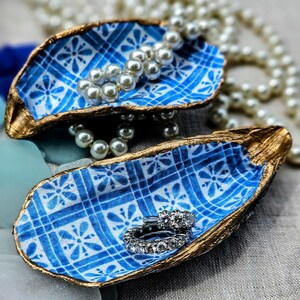 Op de afbeelding: Twee blauwe en witte oesterschelpen met gouden rand. De ene schaal bevat een parelketting en de andere een zilveren ring met diamanten.