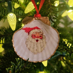 Könnte beinhalten: Ein weißer Muschelschmuck mit goldener Verzierung, der eine Vintage-Illustration von Weihnachtsmann mit roter Mütze und weißem Bart zeigt. Der Schmuck hängt an einem Weihnachtsbaum.