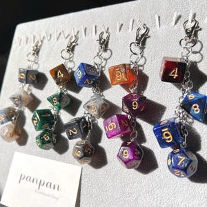 May include: A set of seven colorful dice charms with gold numbers on each side. The dice are attached to silver chains and key rings. The charms are arranged in a row on a white surface. The text "panpan Handmade Shop" is visible in the lower left corner of the image.