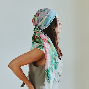 May include: A woman wearing a light blue sleeveless vest with a floral print scarf tied around her head. She is wearing a denim skirt and has her hands in her pockets.