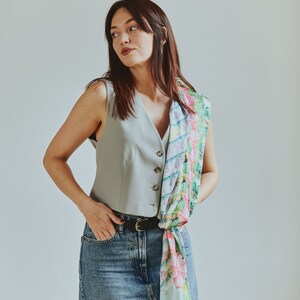 May include: A woman wearing a light blue sleeveless vest with a floral patterned scarf tied around her waist. She is wearing a denim skirt and a black belt.