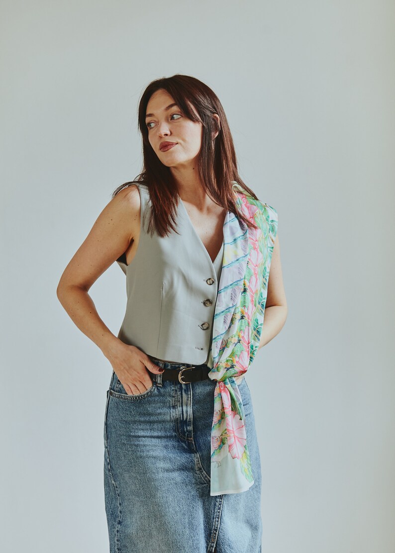 May include: A woman wearing a light blue sleeveless vest with a floral patterned scarf tied around her waist. She is wearing a denim skirt and a black belt.