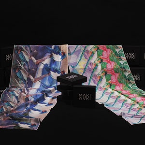 May include: Two silk scarves with different designs, one with a blue bird pattern and the other with a pink flower pattern. The scarves are displayed on a black surface with black boxes that say "Moki" on them.