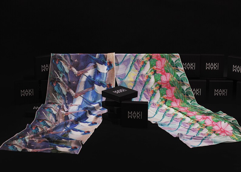 May include: Two silk scarves with different designs, one with a blue bird pattern and the other with a pink flower pattern. The scarves are displayed on a black surface with black boxes that say "Moki" on them.