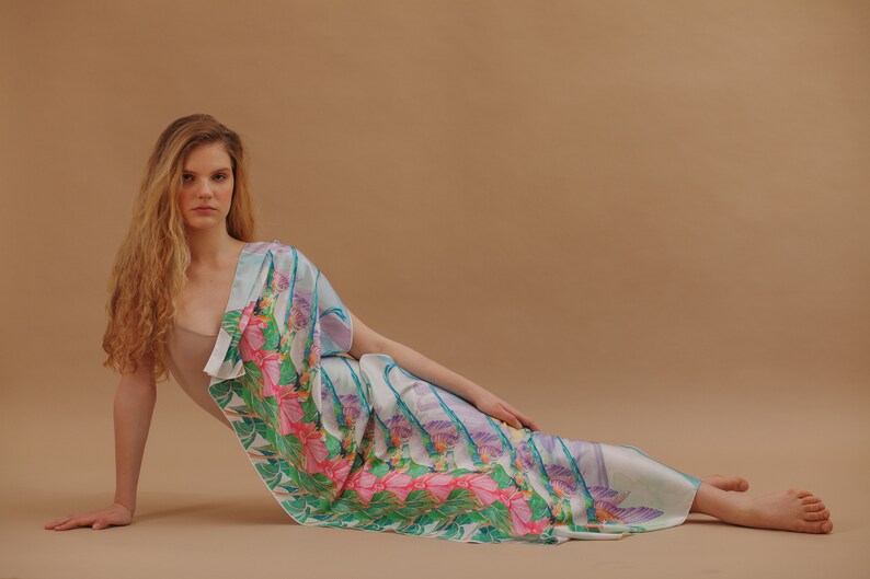 May include: A woman wearing a light blue and white patterned silk scarf with pink and green floral accents. The scarf is draped over her shoulders and around her waist.