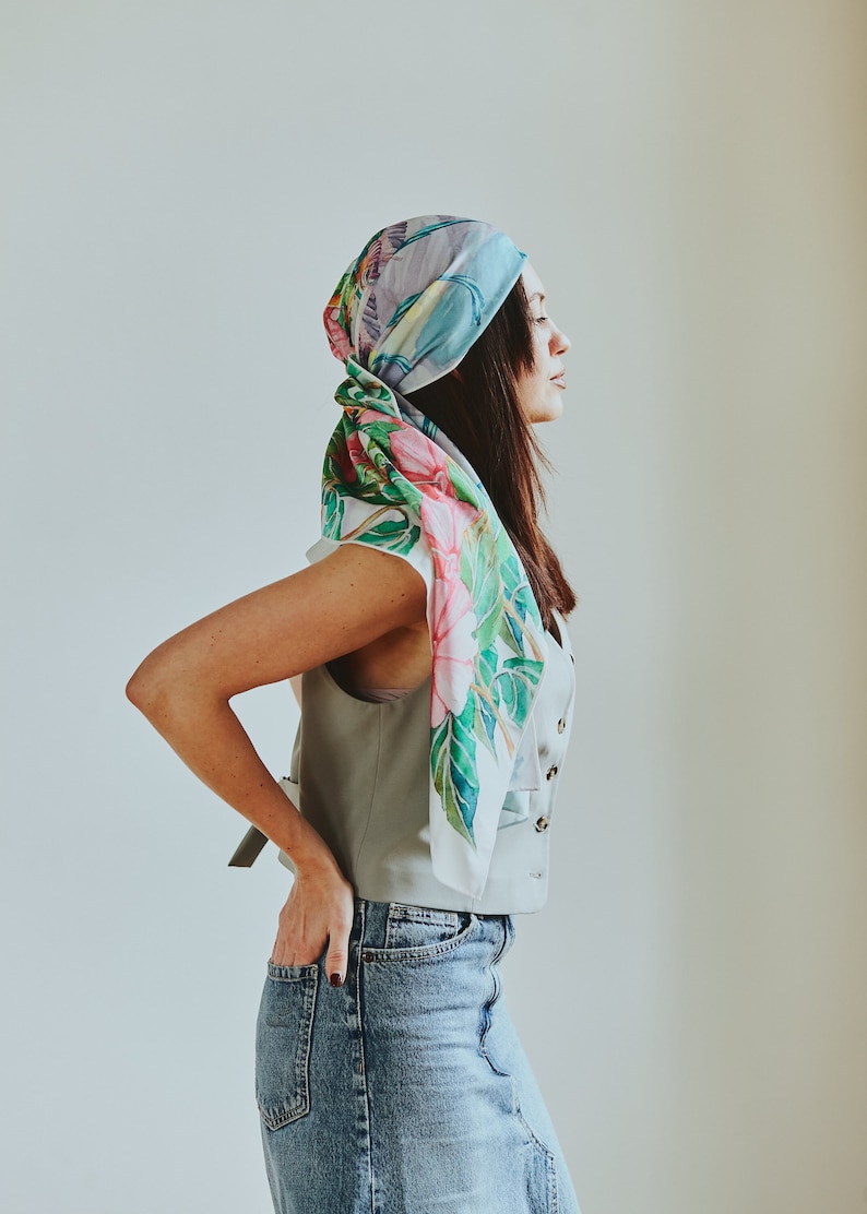 May include: A woman wearing a light blue sleeveless vest with a floral print scarf tied around her head. She is wearing a denim skirt and has her hands in her pockets.