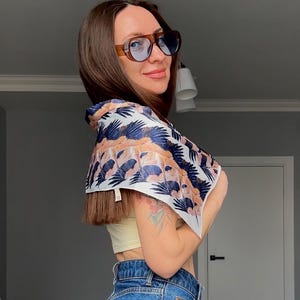 May include: A woman wearing blue jeans and a yellow vest top with a white and blue patterned scarf tied around her neck. She is wearing brown sunglasses.