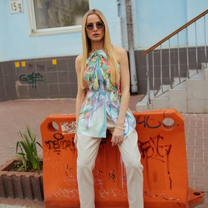 May include: A woman wearing a floral print halter top, beige pants, and black loafers with gold accents. She is standing in front of an orange construction barrier with graffiti.