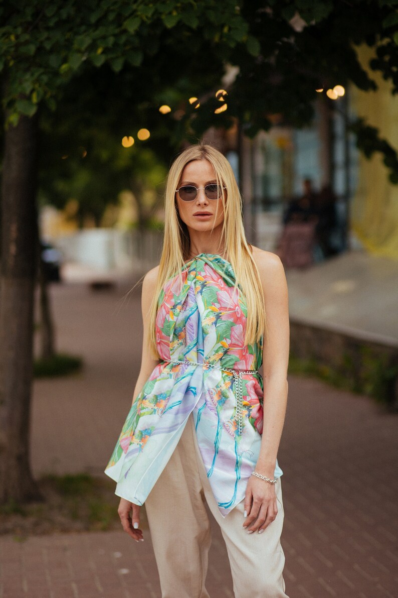 May include: A woman wearing a floral patterned scarf as a top with a silver chain belt and beige pants. She is wearing sunglasses and has long blonde hair.