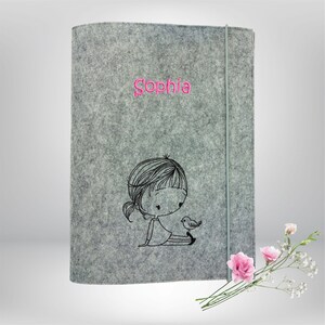 Könnte beinhalten: Graue Filz-Buchhülle mit dem Namen "Sophia" in Pink gestickt. Eine schwarz-weiße Illustration eines Mädchens mit Vogel ist auf der Hülle. Ein graues Gummiband sichert die Hülle.