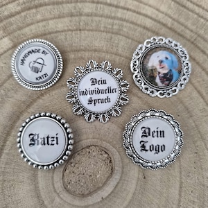 Könnte beinhalten: Eine Sammlung silberfarbener, runder Knöpfe mit verschiedenen Designs. Einige Knöpfe zeigen Texte wie "HANDMADE BY KATZI" und "Katzi", während andere ein Foto eines Hundes und den Text "Dein Logo" und "Dein individueller Spruch" zeigen.