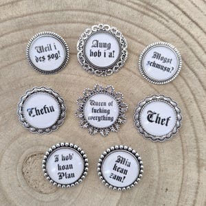 Könnte beinhalten: Sieben silberfarbene Broschen mit weißen Zentren und schwarzem Text. Die Broschen zeigen verschiedene Sprüche, darunter "Queen of fucking everything" und "Chef". Die Broschen haben dekorative Ränder und sind auf einer Holzoberfläche angeordnet.