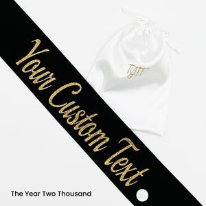 May include: A black sash featuring the gold glitter text Your Custom Text positioned diagonally across the frame, accompanied by a white drawstring pouch with a small gold logo printed on the front surface and the text The Year Two Thousand.
