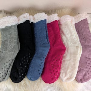 Könnte beinhalten: Sechs Paar gemütliche Slipper-Socken mit Zopfmuster in verschiedenen Farben: Grau, Schwarz, Blau, Fuchsia, Weiß und Lavendel. Jede Socke hat einen flauschigen, weißen Kunstfellbund und rutschfeste Griffe an den Sohlen.