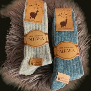 Könnte beinhalten: Zwei Paar Socken aus 100% Alpakawolle. Die Socken sind gerippt und in Grau und Blau erhältlich. Die Verpackung der Socken zeigt ein Lama und den Text "100% Naturfasern mit Alpaca" und "Lieblings-socken".