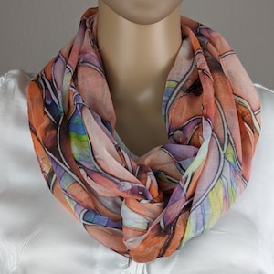 Può includere: Una sciarpa colorata con un motivo astratto in tonalità di arancione, rosa, viola, blu e verde. La sciarpa è realizzata in un tessuto leggero e trasparente ed è annodata in un nodo morbido intorno al collo.