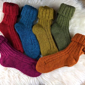 Può includere: Sei paia di calzini colorati a maglia a coste nei toni del rosso, blu, giallo-verde, verde, arancione e viola. I calzini sono ripiegati nella parte superiore e sono disposti su una superficie bianca e soffice.