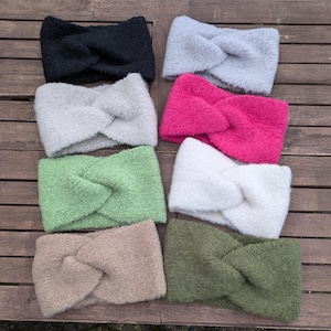 Könnte beinhalten: Acht flauschige Stirnbänder in verschiedenen Farben: Schwarz, Grau, Pink, Grün, Weiß, Beige und Olivgrün. Jedes Stirnband hat ein verdrehtes Knoten-Design. Sie sind auf einer Holzoberfläche angeordnet.
