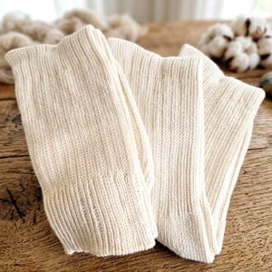 Può includere: Calzini a maglia a coste color crema piegati su una superficie di legno. I calzini sono realizzati in una fibra naturale e morbida. Sullo sfondo, ci sono batuffoli di cotone, filo e piccole pietre.