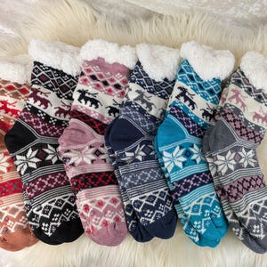 Könnte beinhalten: Sieben Paar warme Socken mit Mustern und weißen Sherpa-gefütterten Bündchen. Die Socken zeigen winterliche Designs wie Schneeflocken und Rentiere in Braun-, Schwarz-, Lila-, Rosa-, Blau- und Grautönen. Sie liegen auf einer flauschigen weißen Oberfläche.