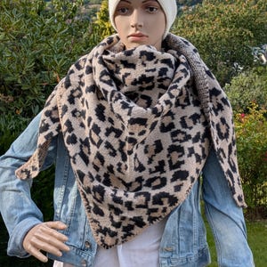 Könnte beinhalten: Ein gestrickter Schal mit Leopardenmuster in Beige und Schwarz. Der Schal ist dreieckig und wird über einer Jeansjacke getragen. Die Mannequin trägt auch eine weiße Mütze.