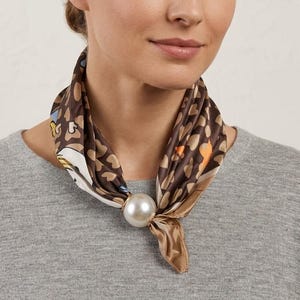 Könnte beinhalten: Ein quadratisches Seidentuch mit braunem Leopardenmuster und Akzenten in Weiß, Blau und Orange, das vorne mit einem großen, runden, weißen Kunstperlenring fixiert ist. Das Tuch ist ordentlich um den Hals über einem grauen Strickpullover gebunden.