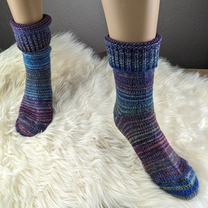 Könnte beinhalten: Ein Paar bunte gestreifte Socken mit umgeschlagenem Bündchen. Die Socken sind aus weichem, gestricktem Stoff und haben verschiedene Blautöne, Lila und Grün.