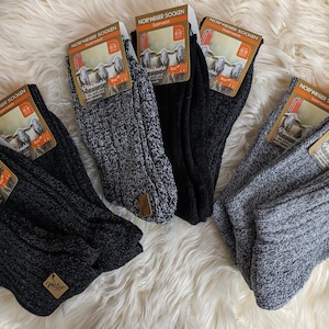 Op de afbeelding: Een stapel van zes paar grijze en zwarte wollen sokken met het label "Norwegier Socken" op elk paar. De sokken zijn gevouwen en gerangschikt op een wit, pluche oppervlak.