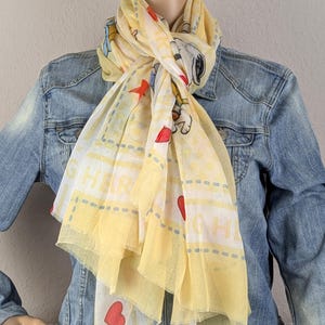 Op de afbeelding: Een gele sjaal met stripfiguren, rode harten en de tekst "HER". De sjaal wordt over een spijkerjasje gedragen. De sjaal heeft een lichte, luchtige textuur.