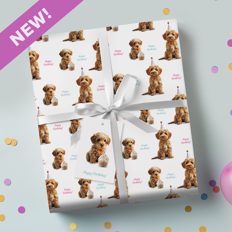 May include: Gift-wrapped present with a white ribbon and tag. The paper displays a repeating pattern of a brown puppy, a cupcake, and the words "Happy Birthday!". A purple banner in the corner reads "NEW!"