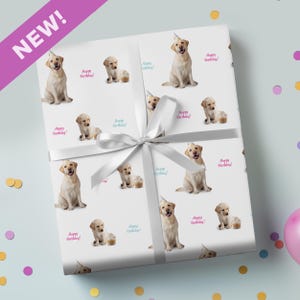 May include: White wrapping paper featuring a repeating pattern of yellow Labrador puppies with party hats and cupcakes. The words "Happy Birthday!" are printed in pink and teal. A white ribbon and bow wrap the gift.